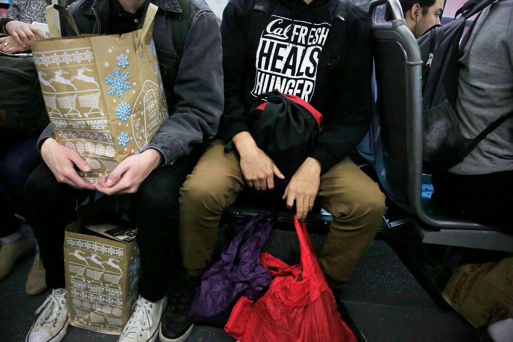 Student wearing "CalFresh Heals Hunger" sweater sits on the bus carrying groceries.