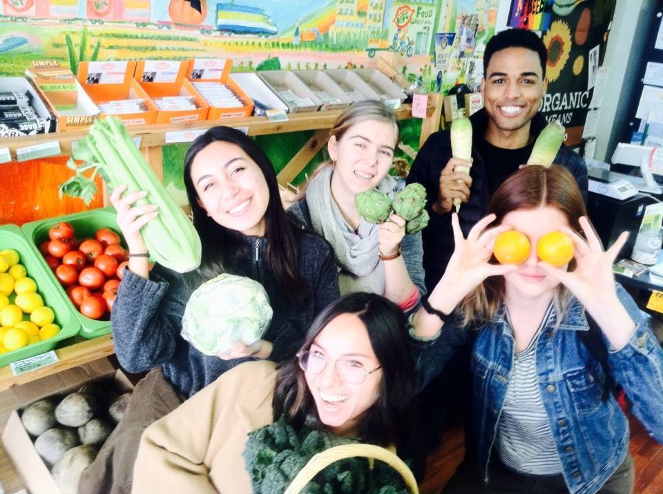 Students holding fresh produce and posing in front of a grocery stand at the Berkeley Student Food Collective.